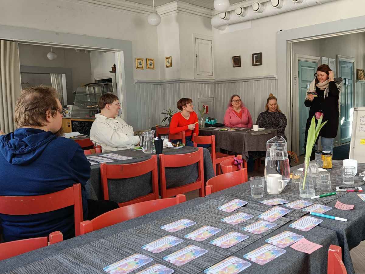 5 ihmistä istuu pöytien ääressä, 1 puhuu ryhmälle katsoen korttia kädessään.