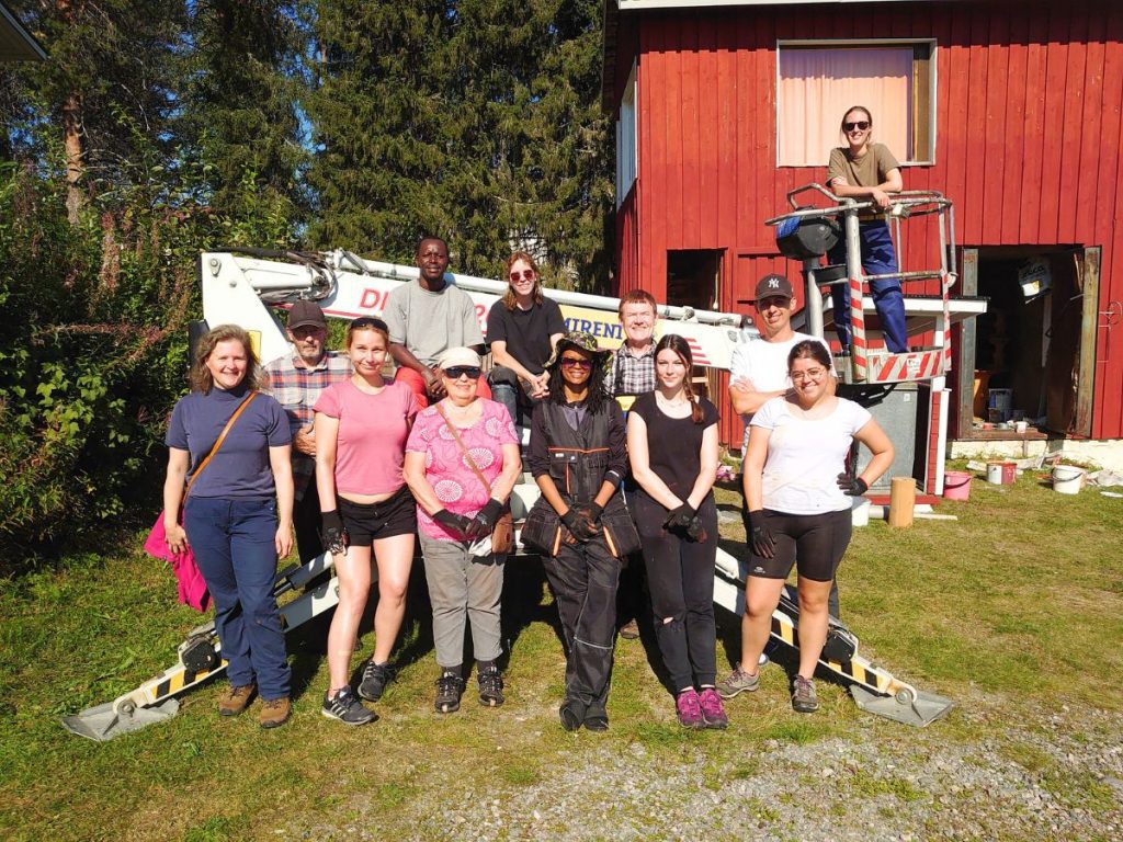 Ryhmä ihmisiä seisoo hymyillen nostokoneen vieressä. Taustalla punaiseksi maalattu rakennus ja puita.