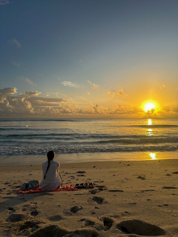 Nainen istumassa rannalla katsoen auringonlaskuun