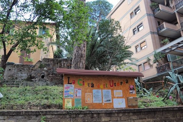 Kyltti, jossa värikkäin kirjaimin teksti 'La Citta dell'Utopia' sekä erivärisiä julisteita. Taustalla kaksi rakennusta ja puita.