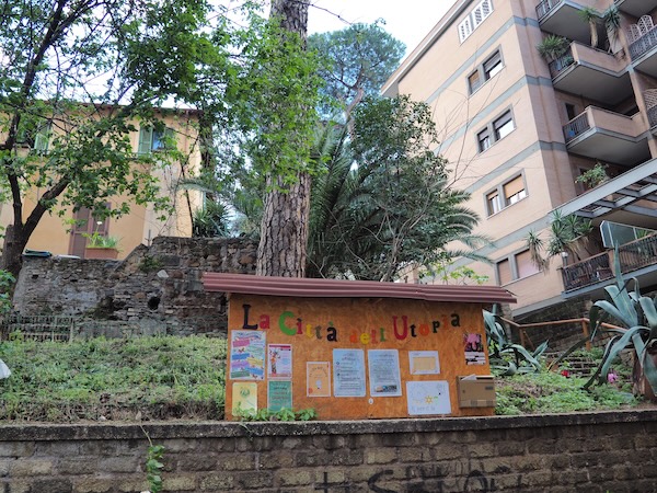 Kyltti, jossa värikkäin kirjaimin teksti 'La Citta dell'Utopia' sekä erivärisiä julisteita. Taustalla kaksi rakennusta ja puita.